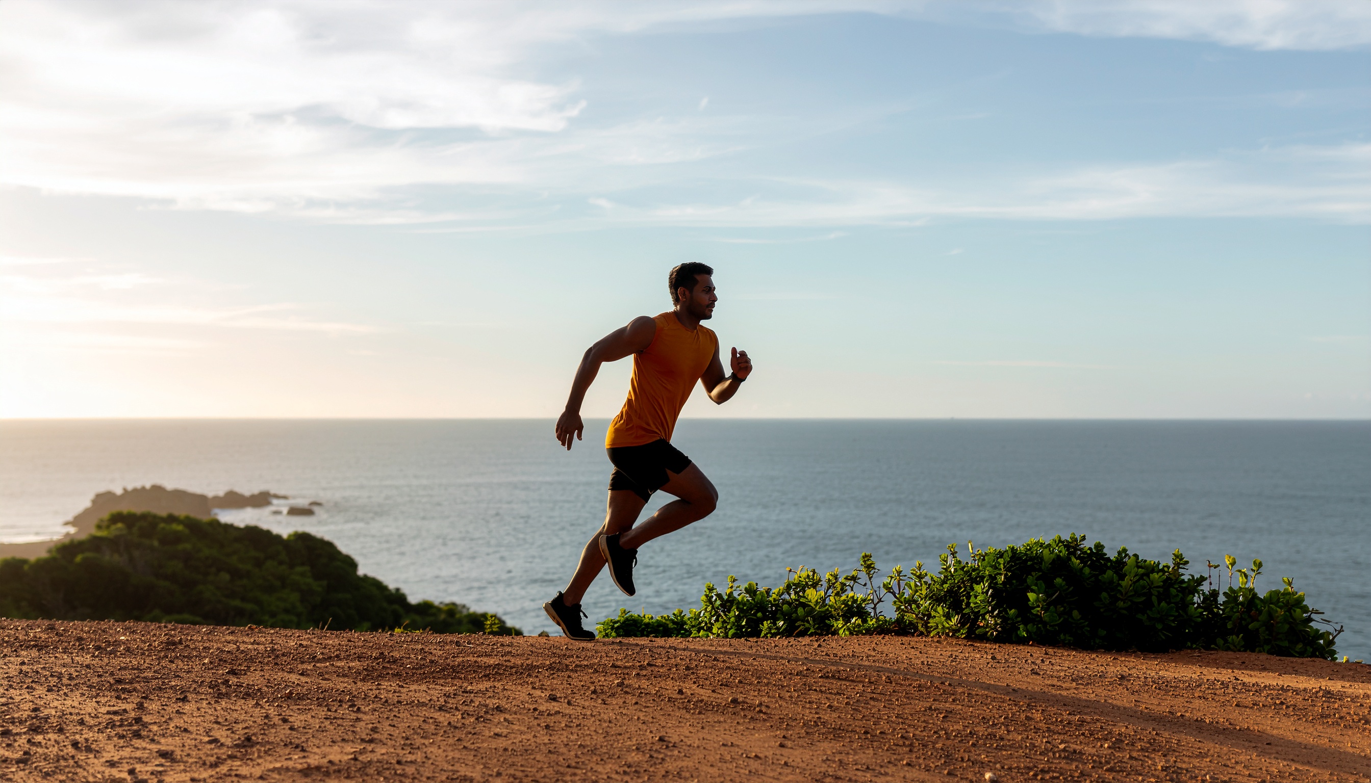 Homem correndo ao ar livre em um trilho de terra, com oceano e céu azul ao fundo. Iluminação suave do pôr do sol cria sombras longas, ressaltando o movimento. A cena transmite energia e liberdade, destacando a conexão com a natureza e o foco na atividade física. Composição equilibrada com contraste entre o solo vermelho e os tons azuis do mar. O ângulo lateral enfatiza a ação e a determinação.